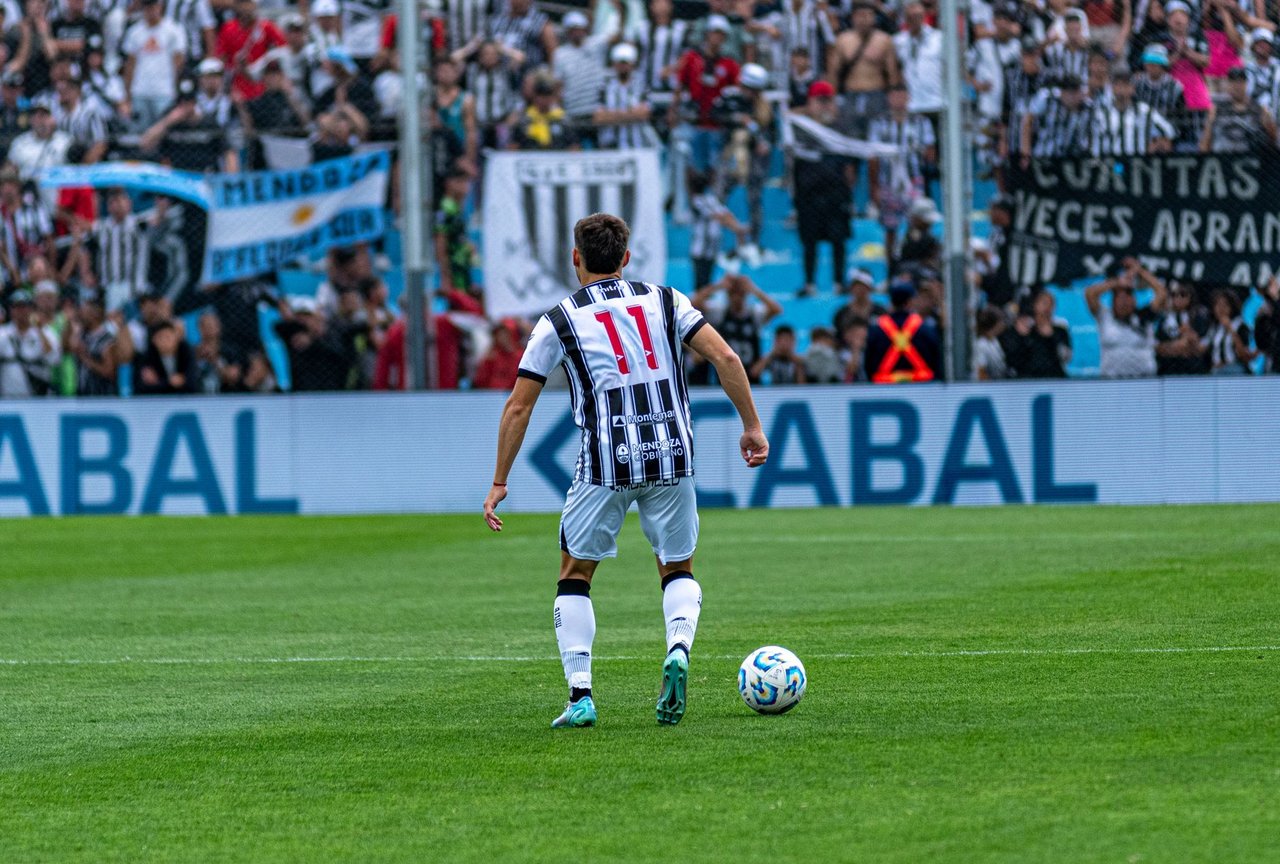 Jogador de futebol com a camisa 11 disputa uma partida diante de torcedores nas arquibancadas.