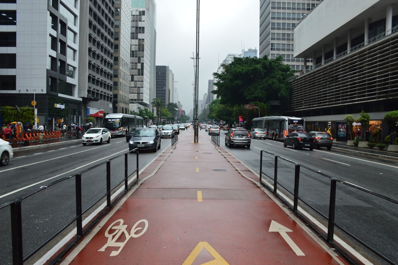 Vista da Avenida Paulista, em São Paulo, com ciclovia, trânsito e edifícios ao redor em um dia nublado.