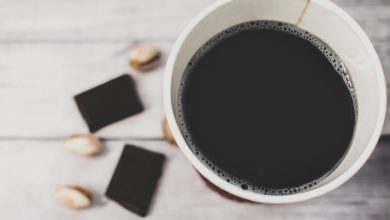 Xícara de café acompanhada de pedaços de chocolate amargo e pistaches sobre uma mesa rústica.