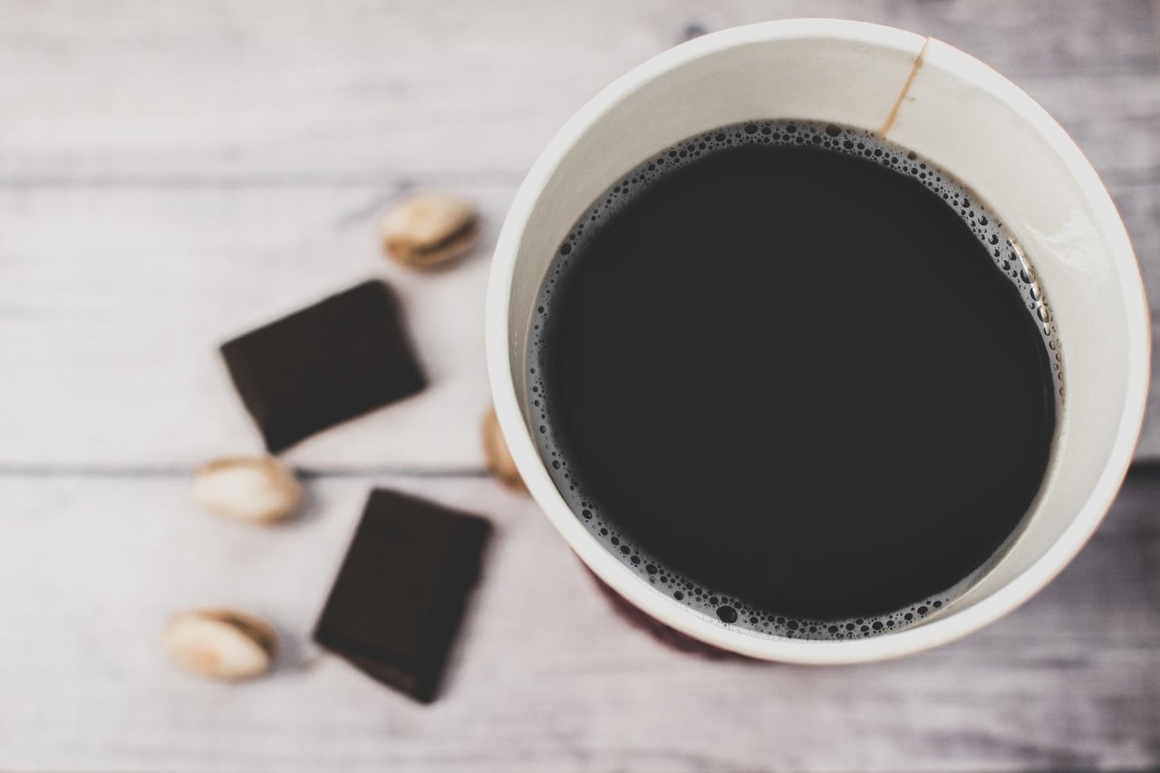Xícara de café acompanhada de pedaços de chocolate amargo e pistaches sobre uma mesa rústica.