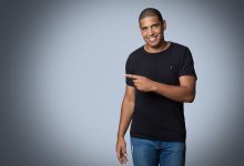 Homem sorrindo, usando camisa preta e calça jeans, aponta para a câmera em um ensaio fotográfico de estúdio.