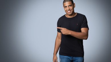 Homem sorrindo, usando camisa preta e calça jeans, aponta para a câmera em um ensaio fotográfico de estúdio.